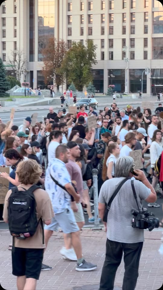 A crowd with raised hands and placards in a city square, with a camera operator in the foreground.