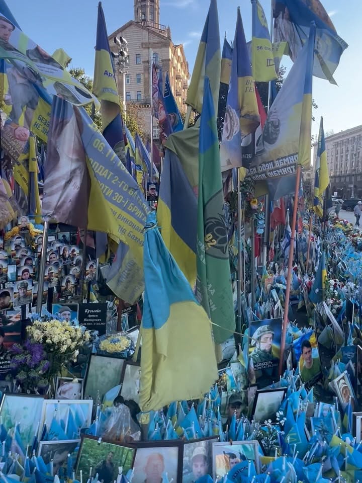 Ukrainian flags bearing portraits rise above photographs and flowers at an outdoor memorial.